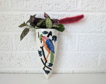 May include: A white ceramic wall vase shaped like a cone with a painted design of a blue and orange bird. The vase is filled with pink and green leafy plants.