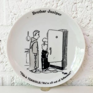 Puede incluir: Plato de cerámica blanca con una caricatura en blanco y negro de dos frailes mirando una nevera. El plato está etiquetado como "Brother Juniper" e incluye el texto "This is TERRIBLE! We're all out of leftovers!"