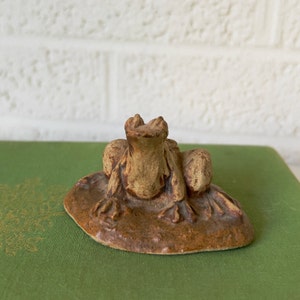 May include: A ceramic frog figurine with a brown and tan glaze, sitting on a green surface. The frog is looking up with its mouth open.