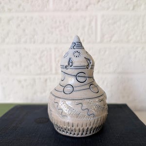 Op de afbeelding: Een beige keramische decoratieve vaas in de vorm van een kalebas met zwarte muzieknoot- en cirkelontwerpen. De vaas heeft een getextureerde basis en een puntige top met een zonontwerp. De vaas staat op een donkerblauwe ondergrond.