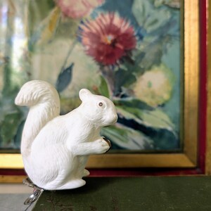 May include: A white squirrel figurine with a bushy tail and a small nut in its paws. The squirrel is attached to a metal clip. The background features a framed floral painting with red and green hues.