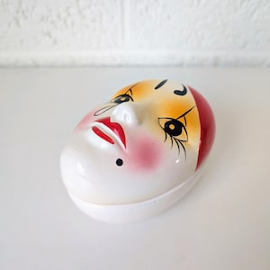 May include: A white, oval-shaped ceramic trinket box with a painted face design. The face features red lips, pink cheeks, black eyelashes, and a red and yellow hair design. A small black dot is below the lip.
