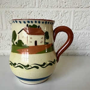 May include: A cream-coloured ceramic jug with a brown handle. The jug has a painted design of a white house with a red roof, green trees, and a brown path. The jug has a blue and green border around the top and bottom.
