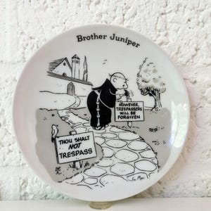 May include: A white ceramic plate featuring a black and white cartoon illustration of Brother Juniper. The plate has the text "Brother Juniper" at the top and a humorous scene with signs that read "Thou Shalt Not Trespass" and "However, Trespassers Will Be Forgiven."