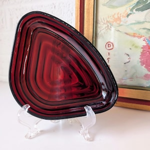 May include: A deep red glass dish with a layered, concentric design. The dish is shaped like a triangle with rounded edges and is displayed on a clear plastic stand.