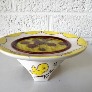 May include: A white ceramic cake stand with a yellow and red rimmed plate. The plate has a yellow center with red dots. The stand has three legs and a yellow bird painted on the front.
