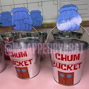 May include: A silver metal bucket featuring the red text CHUM BUCKET and a brown door graphic. A blue mitten cutout is attached to the metal handle. The bucket has a cylindrical shape with a metallic finish and a visible handle for carrying.