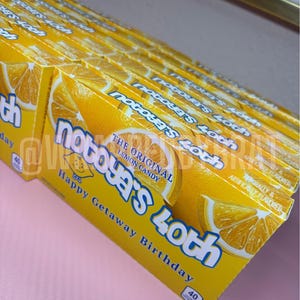 以下が含まれることがあります： レモンのグラフィックと「notoya's 40th Happy Getaway Birthday」の文字が入った黄色のキャンディーボックスの山。箱にはレモンスライスのデザインと「The Original Lemon Candy」の文字が描かれています。