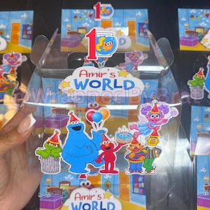 May include: Clear plastic favor boxes decorated with Sesame Street characters, including Cookie Monster, Elmo, and Oscar the Grouch. The boxes feature the text "Amir's World" and a number one, suggesting a first birthday theme.