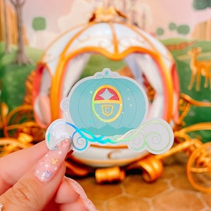 May include: A holographic sticker featuring a teal pumpkin carriage design with gold accents and a red and gold crest. The sticker has a whimsical, fairytale theme, with a dreamy, iridescent finish.