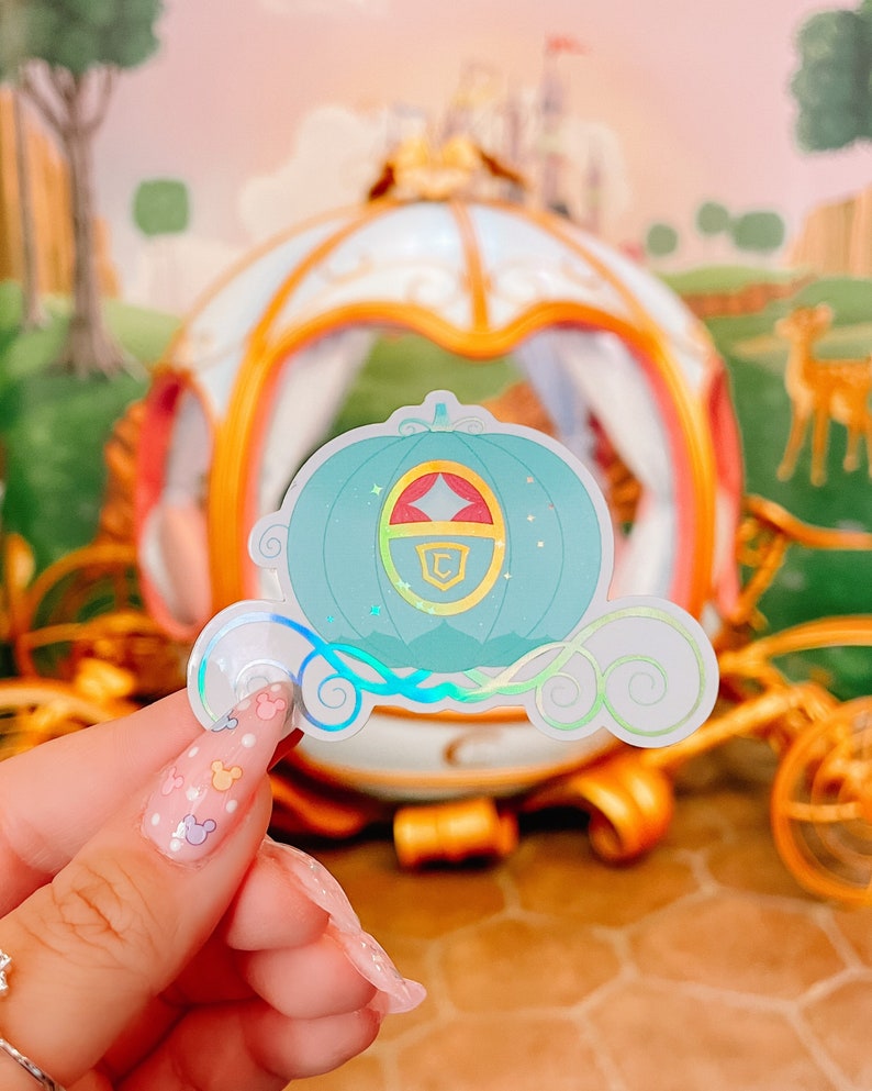 May include: A holographic sticker featuring a teal pumpkin carriage design with gold accents and a red and gold crest. The sticker has a whimsical, fairytale theme, with a dreamy, iridescent finish.