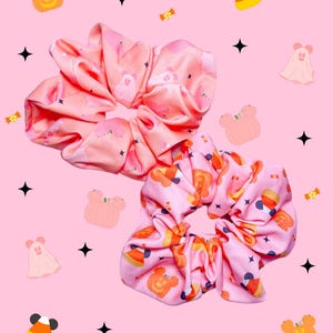May include: Two fabric hair scrunchies featuring a repeating pattern of orange pumpkins, candy corn, and ghost shapes on a light pink background. The scrunchies are gathered with elastic, creating a ruffled, circular shape with a soft texture.