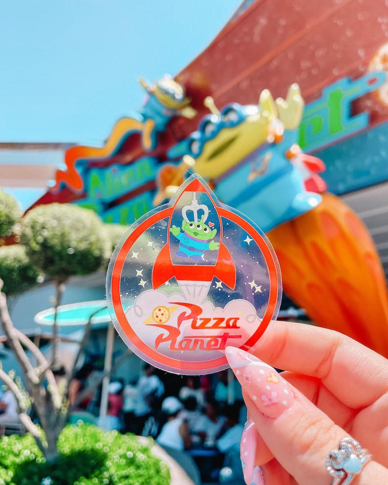 May include: A clear, circular sticker featuring the Pizza Planet logo and a cartoon alien in a rocket ship. The alien is green with three eyes and wearing a blue shirt. The sticker has a red border and a starry background.