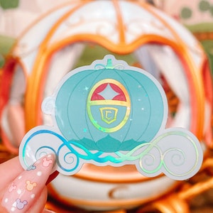 May include: A teal sticker of a fairytale carriage with a gold and red emblem. The sticker has a holographic effect, with swirls and stars. The background shows a blurred image of a carriage.