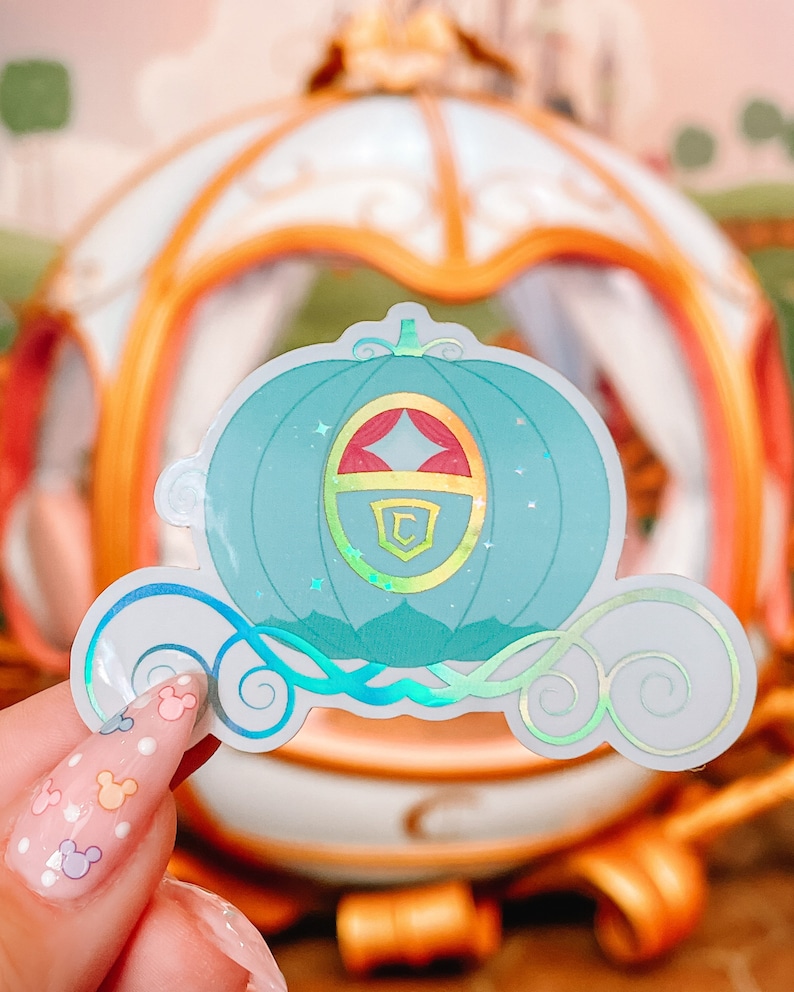 May include: A teal sticker of a fairytale carriage with a gold and red emblem. The sticker has a holographic effect, with swirls and stars. The background shows a blurred image of a carriage.