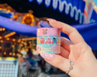 以下が含まれることがあります： ディズニーのキャラクターとディズニーランドのロゴが入った装飾用マスキングテープのロールのスタック。 ロールはピンク、青、緑などのパステルカラーです。 テープは指輪をした手で持たれています。