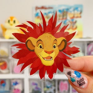 May include: A clear sticker featuring a cartoon lion's face with a red mane. The lion has a yellow face, brown ears, and a smiling expression. The sticker is held up by a hand with decorated nails. Other stickers are visible in the background.