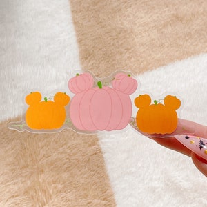 May include: A clear sticker featuring three pumpkin shapes with rounded ears. The center pumpkin is light pink, while the two flanking pumpkins are orange. Each pumpkin has a small green stem and thin green vine details on a transparent background.