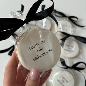 May include: Round cookies with white icing and the text "I can't say I do without you" are individually wrapped and tied with black satin ribbon. The cookies are a light beige colour and are packaged in clear cellophane.