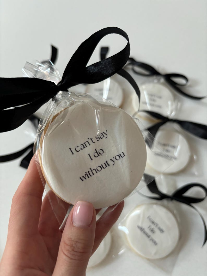 May include: Round cookies with white icing and the text "I can't say I do without you" are individually wrapped and tied with black satin ribbon. The cookies are a light beige colour and are packaged in clear cellophane.