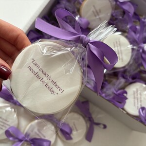 May include: Round cookies individually wrapped in clear cellophane and tied with purple satin ribbon. Each cookie has the phrase "I am exactly where I need to be today." printed on it. The cookies are in a white box.
