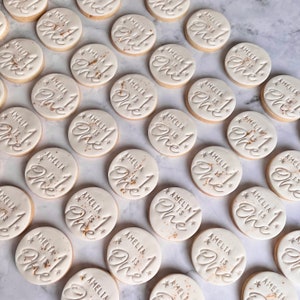 May include: A selection of round biscuits with white icing. Each biscuit is decorated with the text "AMELIA is One!" in a stylish script, with star details and gold flecks. The biscuits are arranged on a light-coloured surface.