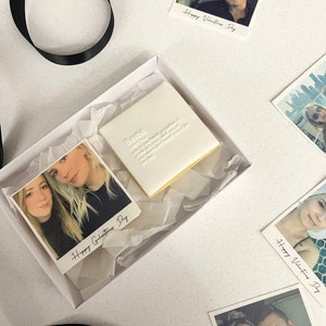 May include: A white gift box contains a white cookie with the word "Bestie" and definition, and several small photo prints. The prints have photos of people and the text "Happy Valentines Day" or "Happy Galantines Day". A black ribbon is also present.