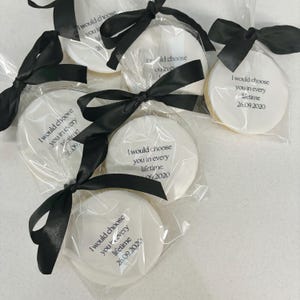 May include: Round white cookies in clear cellophane, tied with black satin ribbons. Each cookie is printed with the text "I would choose you in every lifetime" and a date. A delightful treat for a celebration.