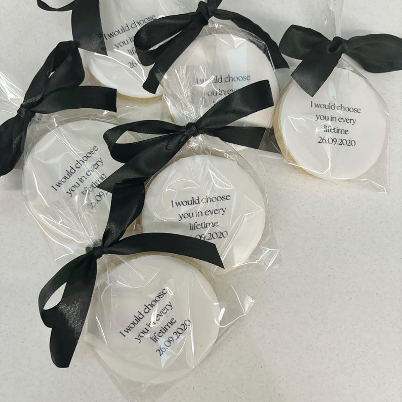 May include: Round white cookies in clear cellophane, tied with black satin ribbons. Each cookie is printed with the text "I would choose you in every lifetime" and a date. A delightful treat for a celebration.