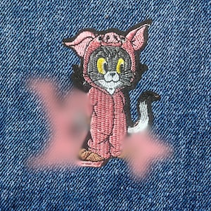 May include: Embroidered patch featuring a cartoon cat in a pink costume, set against a blue denim background. The cat has yellow eyes and a white face. The denim has a frayed edge.