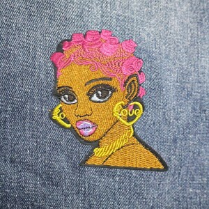 May include: Embroidered patch featuring a stylized woman with pink hair and gold jewelry. The patch has a black border and is set against a denim background. The earrings spell out the word "love".
