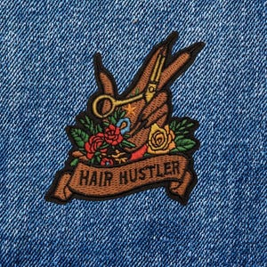 May include: Embroidered patch featuring a hand holding scissors, surrounded by roses and a banner that reads "HAIR HUSTLER." The patch is brown, with gold scissors and colorful floral accents, set against a blue denim background.