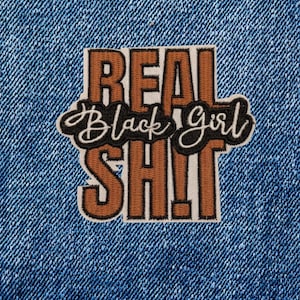 May include: Close-up of a blue denim fabric with a frayed edge. A rectangular patch is sewn onto the denim, featuring the words "REAL Black Girl SH!T" in brown and black lettering.