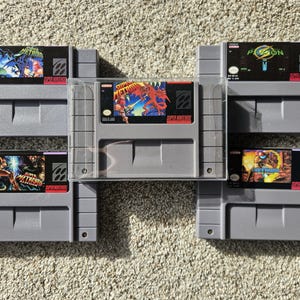 Puede incluir: Colección de cinco cartuchos de juegos de Super Nintendo Entertainment System (SNES). Los cartuchos grises presentan etiquetas de juegos coloridas con títulos como "Super Metroid" y "Palson". Los juegos están dispuestos sobre una superficie marrón claro.