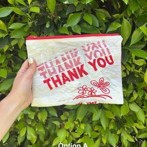 May include: Cream-colored zippered pouch with red text that reads "THANK YOU" repeated multiple times. The pouch has a red zipper and a red floral design. The background is a lush green bush.
