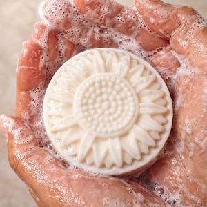 May include: A round, off-white bar of soap featuring a molded sunflower design on its surface. The soap is covered in a thick, bubbly white lather, highlighting the intricate petal and seed details embossed into the center of the circular bar.