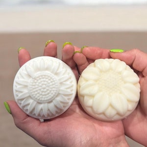 May include: Two white soap bars shaped like sunflowers. The soap bars are in the palm of a person's hand.