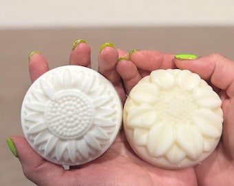 May include: Two white soap bars shaped like sunflowers. The soap bars are in the palm of a person's hand.