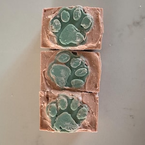 May include: Three bars of soap with a paw print design. The soap is a light pink color with a green paw print.