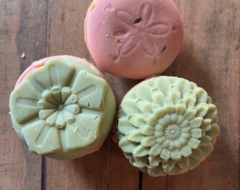 May include: Three decorative soaps. One is light pink with a sand dollar design, and the other two are green with floral patterns. The soaps are round and sit on a wooden surface.