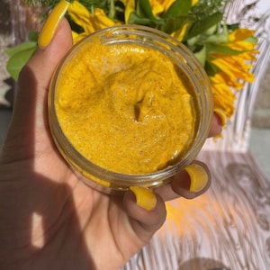 Puede incluir: Primer plano de un pequeño frasco transparente lleno de una mascarilla facial espesa de color amarillo anaranjado. La mascarilla tiene una textura suave y cremosa.