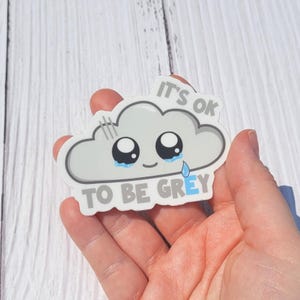 May include: A die-cut sticker shaped like a light grey cloud with a smiling face, large black eyes, and blue teardrops. The text IT'S OK TO BE GREY is printed in grey and blue block letters above and below the cloud character on a white background.