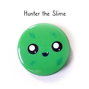 May include: A round green pin-back button featuring the text Hunter the Slime. The button displays a cartoon face with large black eyes, white highlights, and a small smiling mouth, surrounded by four small dark green insect illustrations.