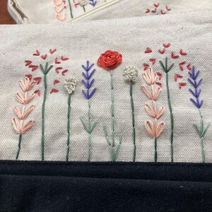 May include: Two off-white canvas pouches with embroidered floral designs. The embroidery features red, pink, purple, and white flowers with green stems. The bottom of the image shows a black fabric.
