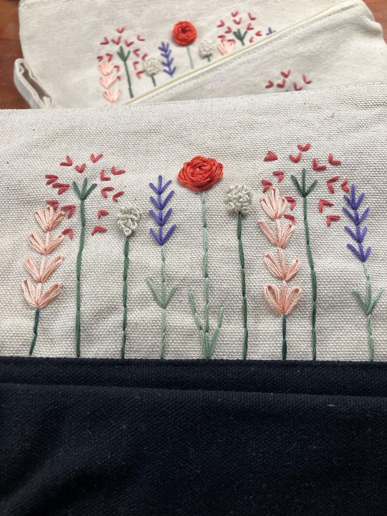 May include: Two off-white canvas pouches with embroidered floral designs. The embroidery features red, pink, purple, and white flowers with green stems. The bottom of the image shows a black fabric.