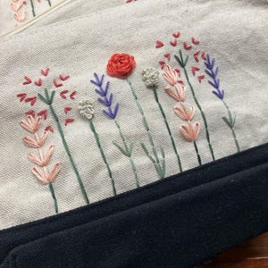 May include: Close-up of two canvas pouches with embroidered floral designs. The pouches feature a cream-colored fabric with a black bottom. The embroidery includes red roses, lavender, and other colorful flowers.