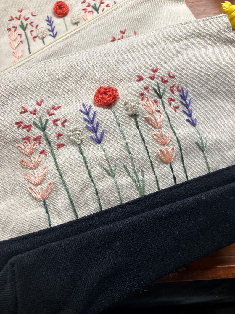 May include: Close-up of two canvas pouches with embroidered floral designs. The pouches feature a cream-colored fabric with a black bottom. The embroidery includes red roses, lavender, and other colorful flowers.