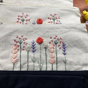 May include: Three canvas pouches with embroidered floral designs. The pouches have a natural beige color with a black base. The embroidery features red, pink, purple, and white flowers with green stems. Each pouch has a zipper closure.