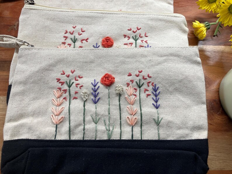 May include: Three canvas pouches with embroidered floral designs. The pouches have a natural beige color with a black base. The embroidery features red, pink, purple, and white flowers with green stems. Each pouch has a zipper closure.