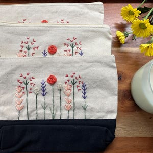 May include: Three canvas zipper pouches with embroidered floral designs. The pouches have a cream-colored body with black bottoms and feature red, pink, and purple flower embroidery. A candle and yellow flowers are in the background.